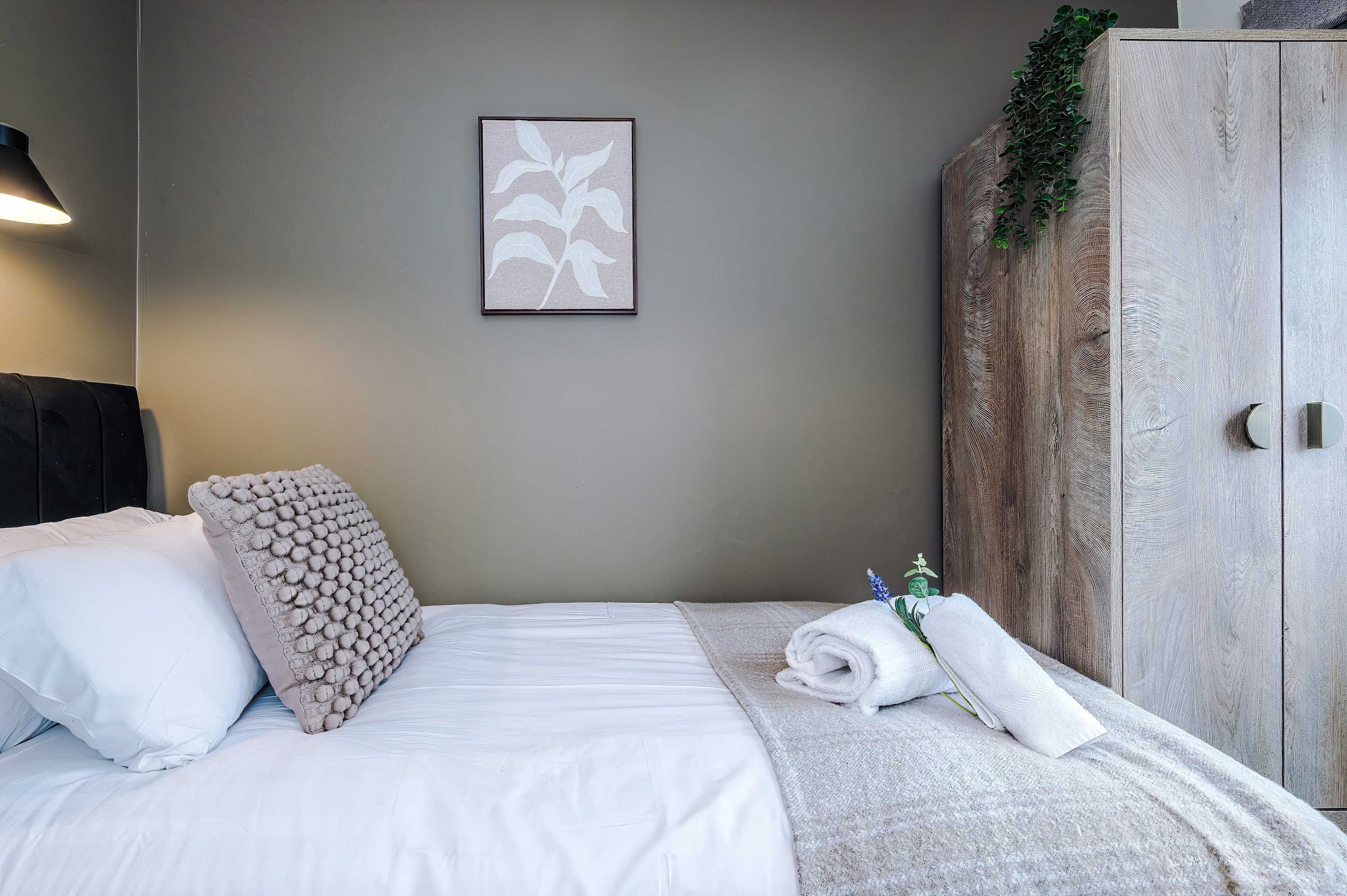 Un lit bien fait, avec des draps blancs, un coussin décoratif et une serviette enroulée. Une œuvre d’art encadrée est accrochée au mur au-dessus du lit.