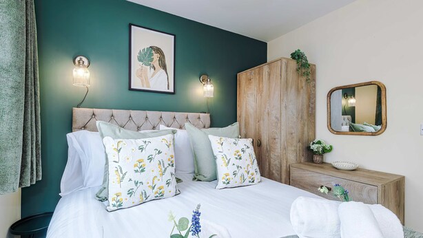 Ein ordentlich bezogenes Bett mit floralen Kissen, ein Holzschrank, ein Nachttisch mit Pflanze und ein an der Wand befestigter Spiegel.