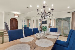 A dining area with a wooden table, blue upholstered chairs, a chandelier, and a television in the background. - 6 Bedroom Home with Pool 10 minute Walk To Beach (Miramar Beach)
