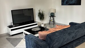 Un soggiorno moderno con una televisione a schermo piatto, un divano blu scuro, un tavolino da caffè, una lampada da terra e un quadro appeso al muro.