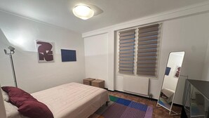 Una camera da letto con un letto, una lampada da terra, un termosifone, una finestra con persiane, uno specchio e un tavolino.