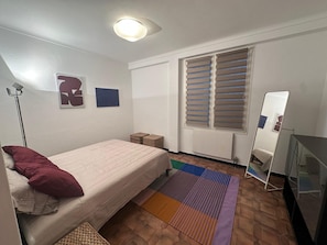 Ein Schlafzimmer mit Bett, Stehlampe, Heizkörper, Fenster mit Jalousien, Spiegel und einem kleinen Tisch.
