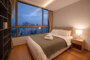 Ein Hotelzimmer mit großem Fenster, einem Bett mit weißer Bettwäsche, einem Nachttisch mit Lampe und einem Bücherregal.
