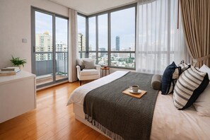 Ein modernes Schlafzimmer mit einem großen Bett, einem Sessel, einem Schreibtisch und Blick auf die Stadt durch große Fenster.