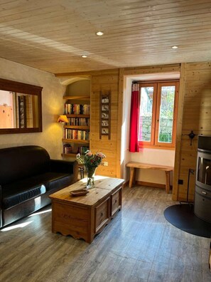 A cozy room with a wooden ceiling, a black leather sofa, a wooden coffee table with a vase of flowers, a bookshelf, a window with red curtains, and a stove. - Fireplace or stove, parking, tv, 40m², Peisey-Vallandry (Peisey-Nancroix)