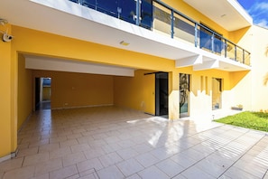 A modern building with a yellow facade, a covered outdoor area with tiled flooring, and a glass balcony.