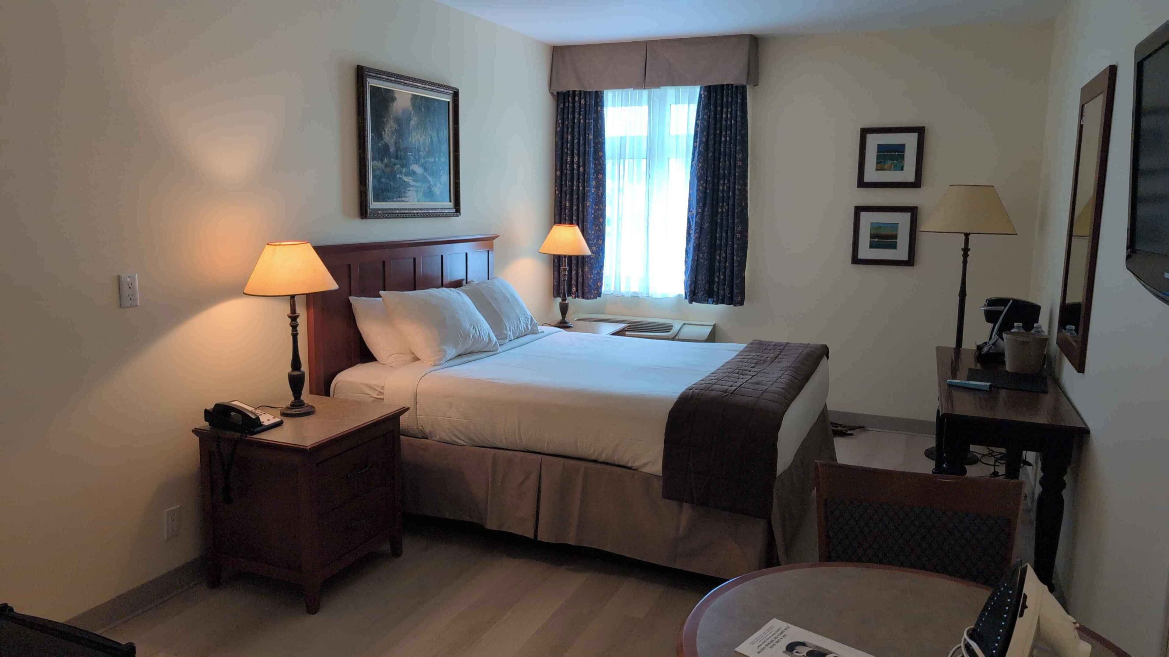 Ein Hotelzimmer mit Bett, zwei Nachttischlampen, einem Nachttisch, einem Schreibtisch, einem Sessel und einem Fenster mit Vorhängen, sowie einem Bild an der Wand.