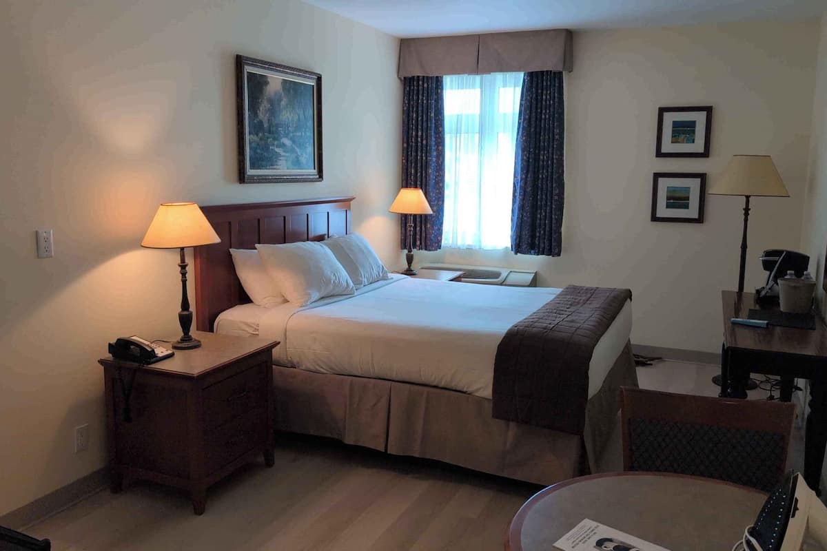 A hotel room with a bed, two bedside lamps, a nightstand, a desk, a chair, a window with curtains, and a painting on the wall.