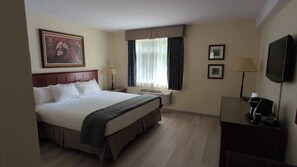Ein Hotelzimmer mit einem Bett, einem Schreibtisch, einem Sessel, einem Fernseher, einem Fenster mit Vorhängen und einem an der Wand hängenden Bild.