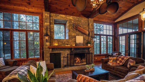 L’intérieur chaleureux d’un chalet avec une cheminée en pierre, des murs en bois et de grandes fenêtres offrant une vue sur la forêt.