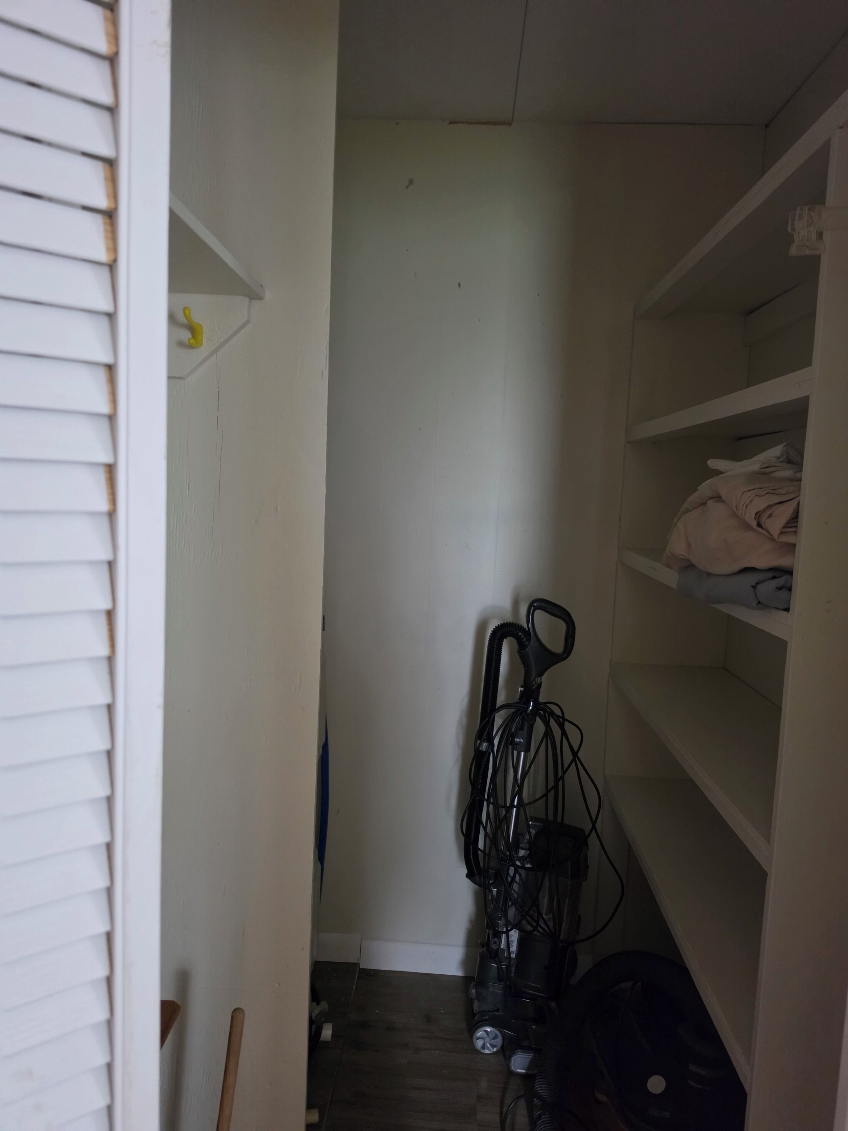 A narrow hallway with a white door on the left, a vacuum cleaner in the center, and a shelf with folded clothes on the right.