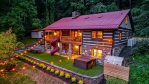 Eine Blockhütte mit rotem Dach, einem Steinkamin und einer Holzterrasse mit Außenbeleuchtung.