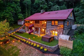 A log cabin with a red roof, a stone chimney, and a wooden deck with outdoor lighting. - Log Cabin, Theater, Arcade & Sauna | Beaver Cabin (Weaverville)