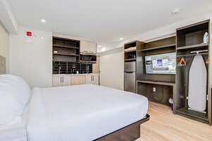 Quarto de hotel com cama, cozinha compacta, televisão e espaço para guardar pertences.