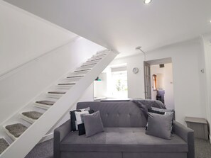 Um interior moderno com um sofá cinzento, uma escadaria branca e uma porta que dá para outro quarto.