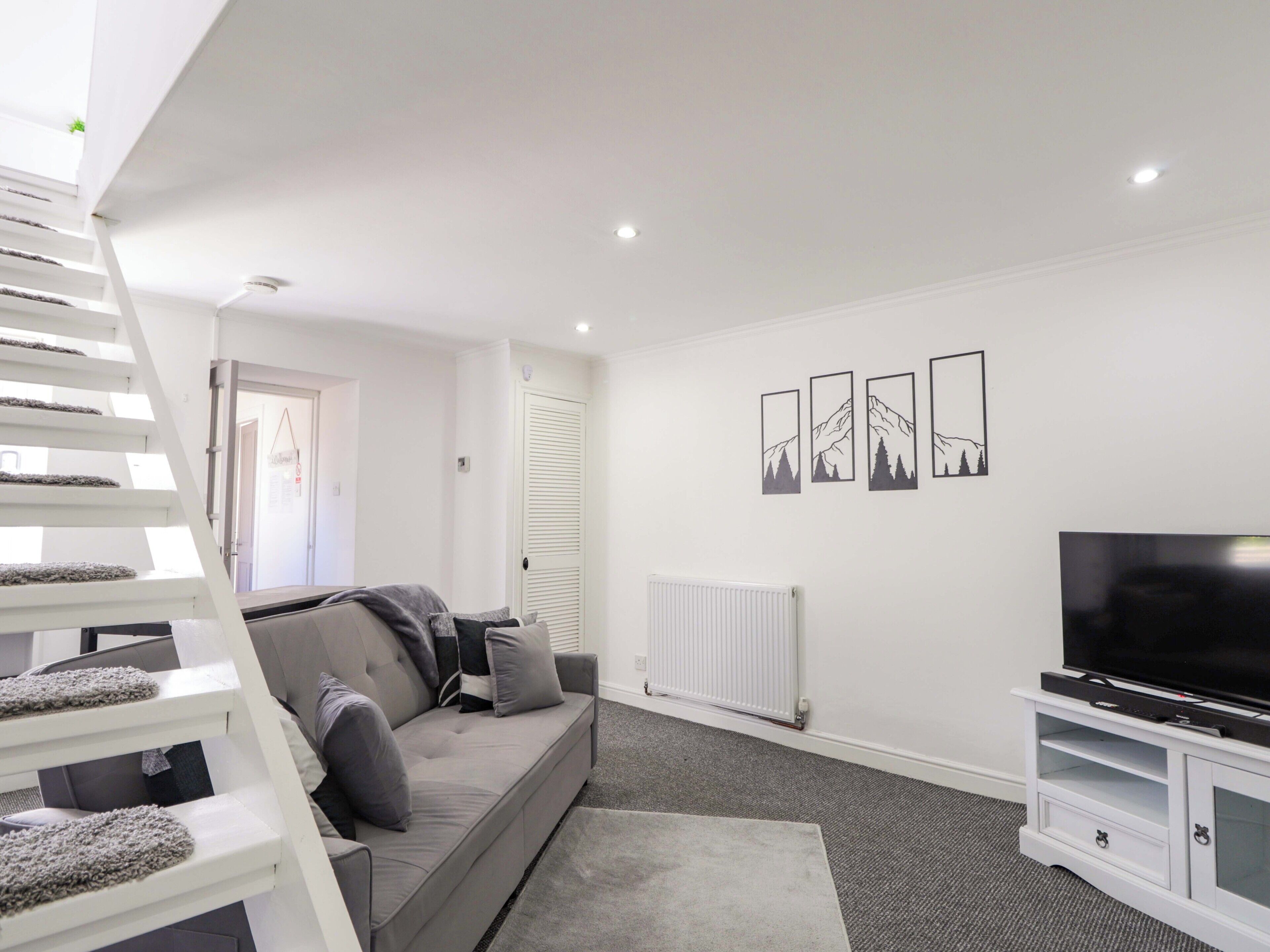 A modern living room with a grey sofa, a white entertainment unit, a TV, a radiator, and a staircase.