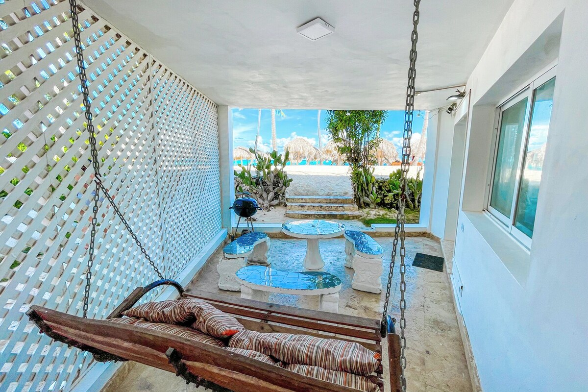 A room with a swing, a small table, a chair, and a view of the beach.