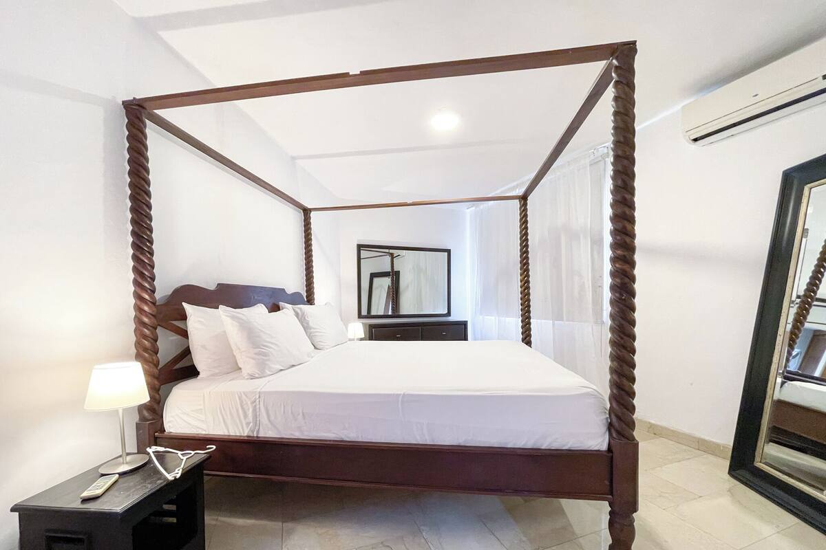 A four-poster bed with white linens, a bedside table with a lamp, and a mirror on the wall.