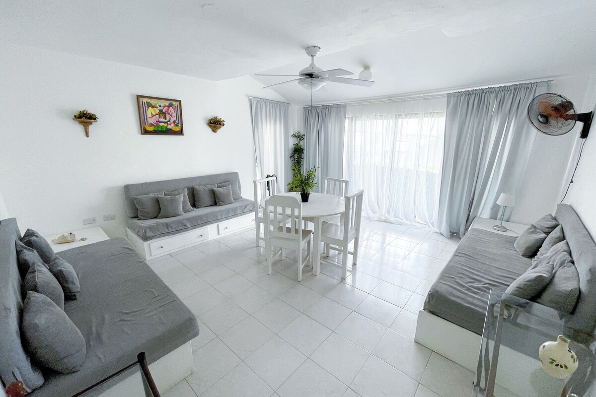A modern living room with a sofa, dining table, and chairs, a ceiling fan, and a large window with curtains.