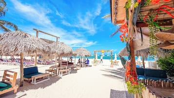 A beach setting with thatched umbrellas, wooden lounge chairs, and a sandy beach.