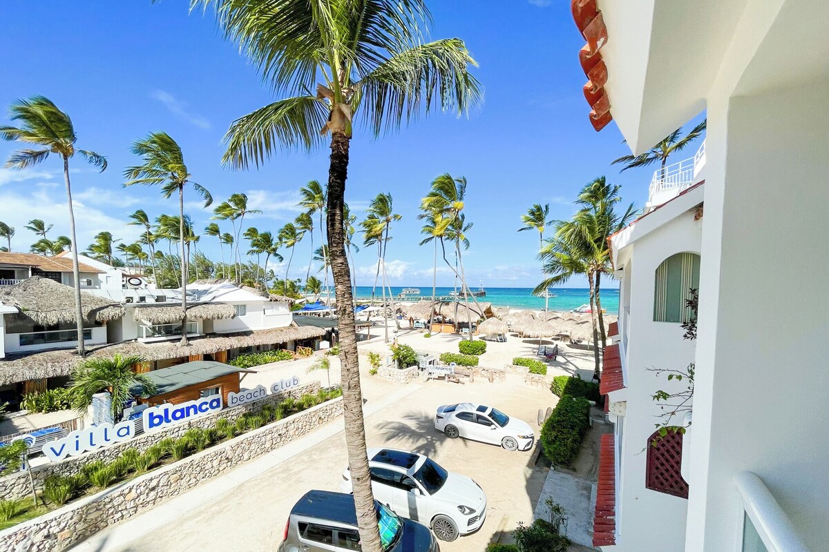 A beach view with palm trees, a beach club sign, and parked cars.
