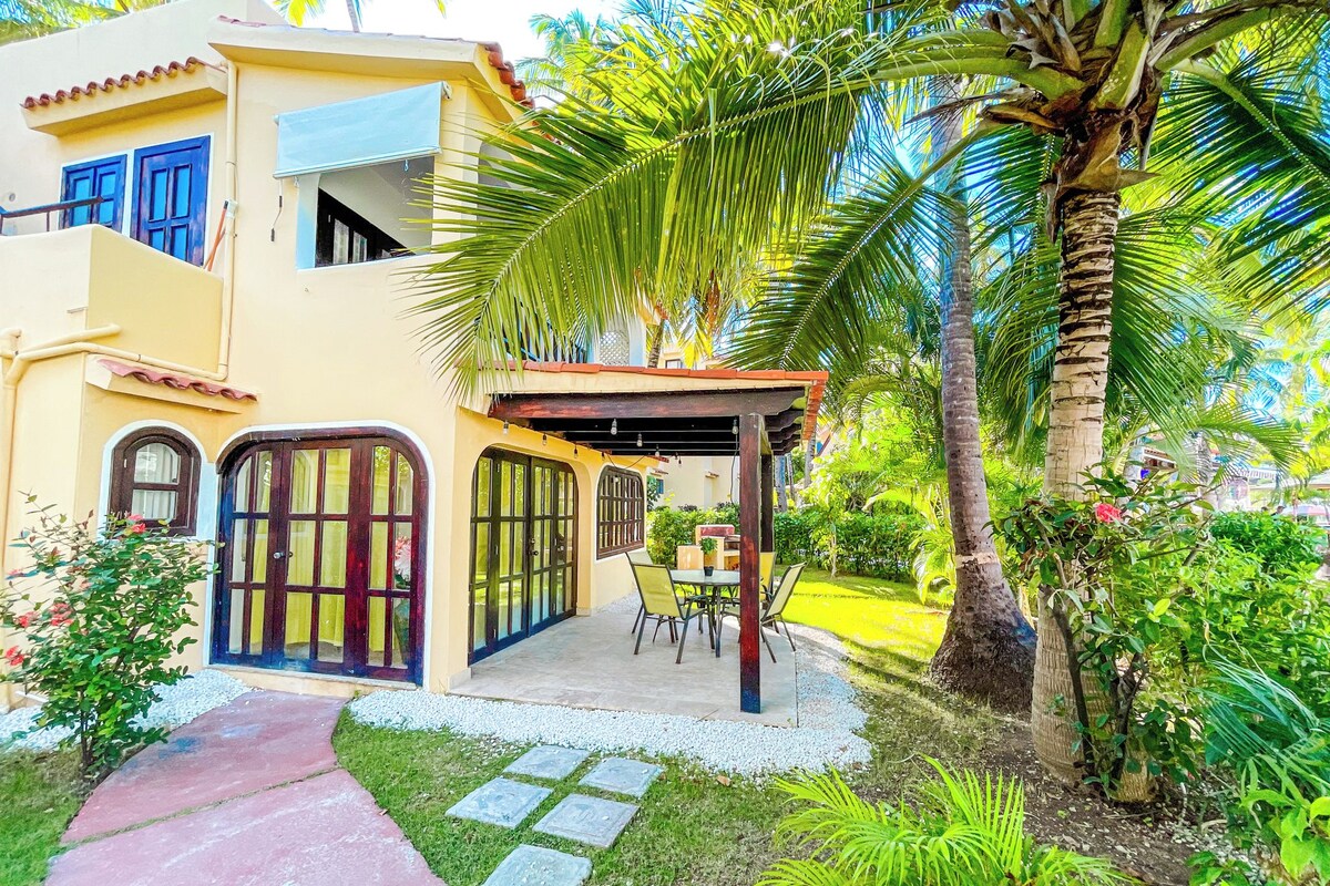 Un edificio a due piani con un portico coperto, una palma e un giardino con lastre di pietra.