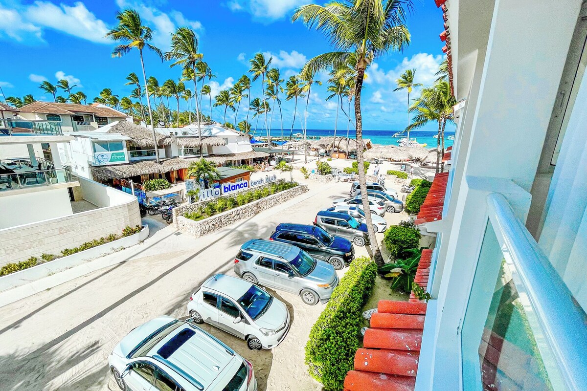 A beachside view with parked cars, a beach club, and palm trees.