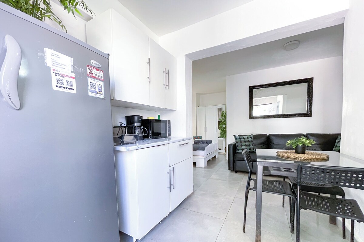A compact kitchen with a refrigerator, microwave, and coffee maker. There is a dining area with a table and chairs, and a sofa in the background.