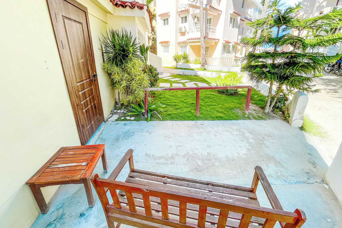 Un balcone con una panca e un tavolo in legno, un piccolo giardino con piante e vista sugli edifici circostanti.