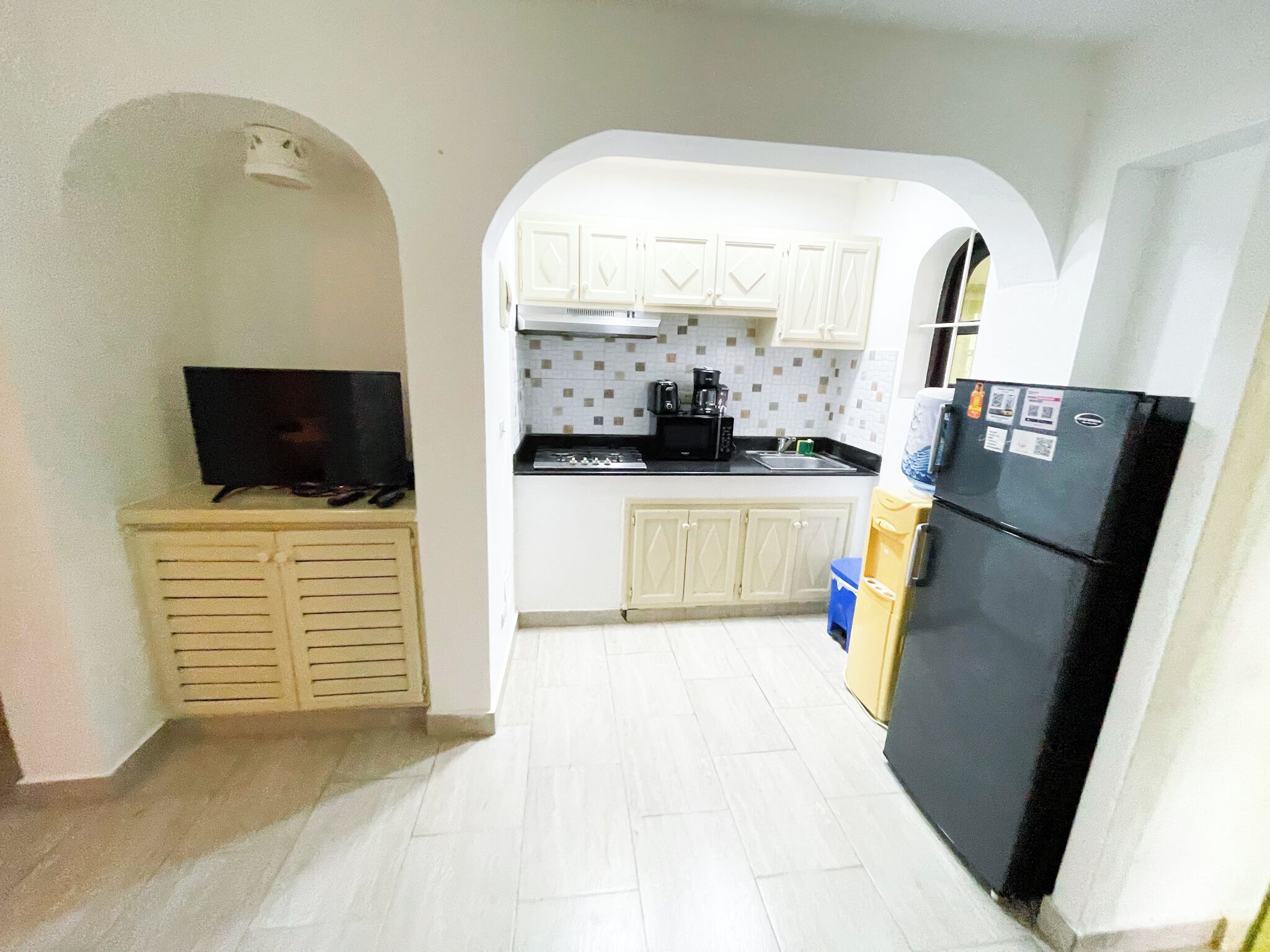 Cocina compacta con estufa, microondas y refrigerador, gabinetes blancos y un televisor sobre un mueble.