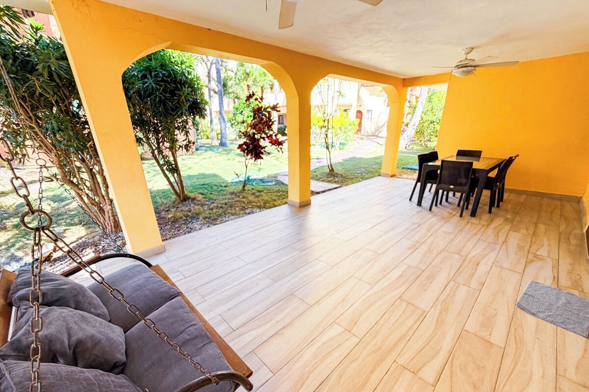 Un portico coperto con una sedia a dondolo, un tavolo da pranzo con sedie e vista su un giardino.
