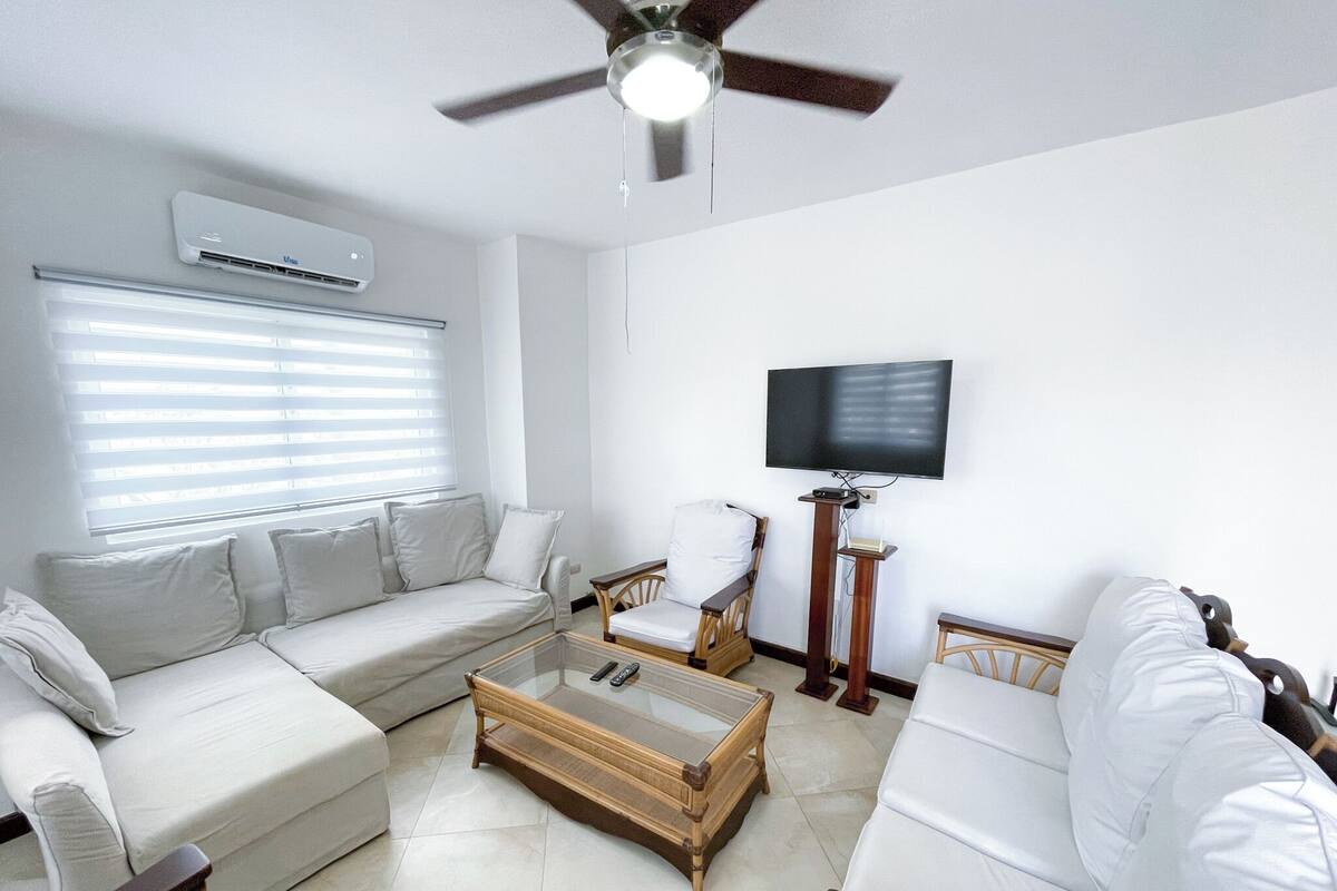 A living room with a sofa set, a coffee table, a television mounted on the wall, and a ceiling fan.
