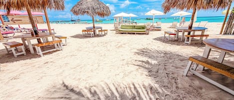 A beach with thatched umbrellas, wooden tables, and lounge chairs.