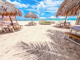 A beach with thatched umbrellas, wooden tables, and lounge chairs.