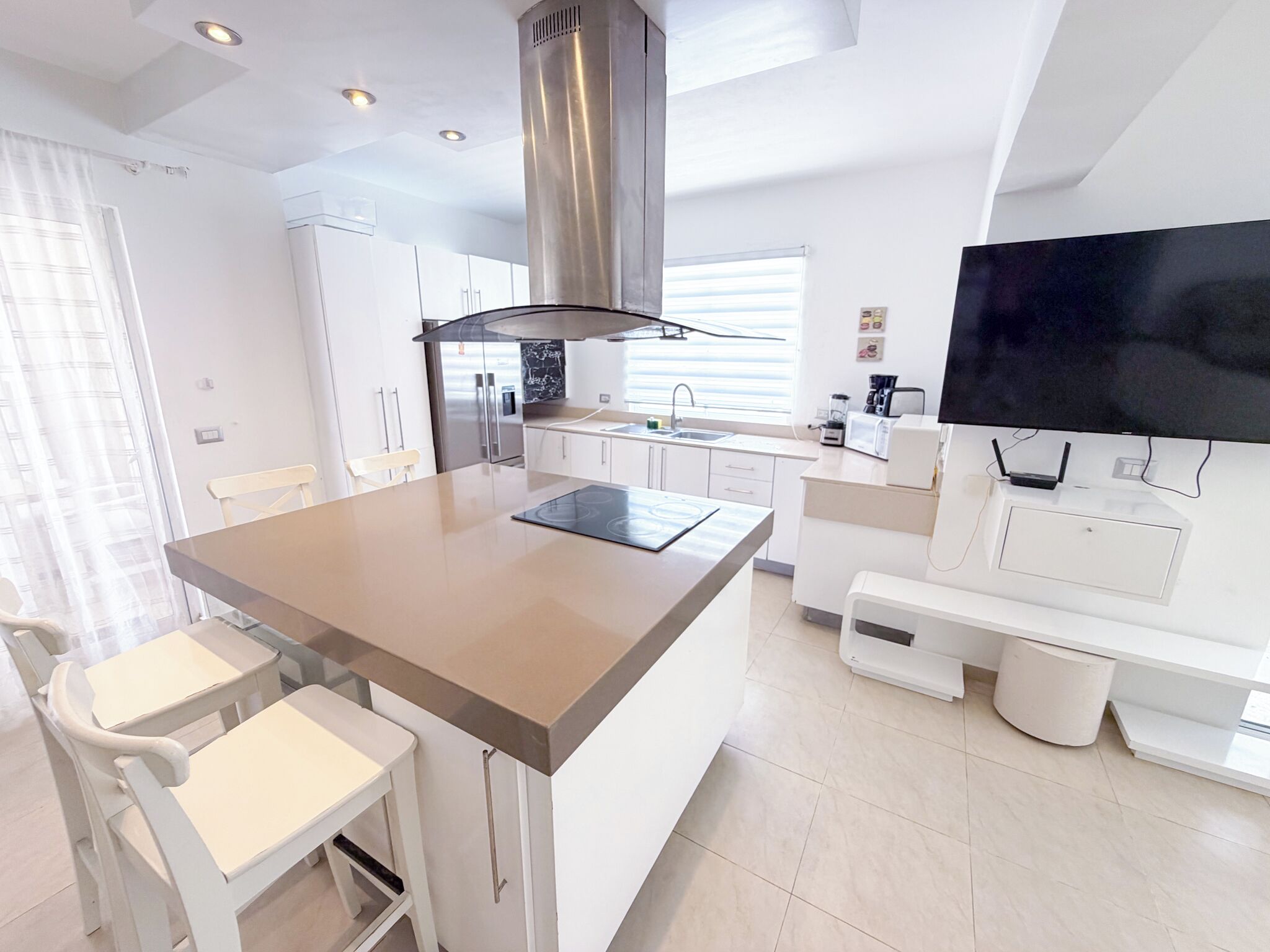 A modern kitchen with a central island, white cabinetry, a stainless steel range hood, a built-in TV, and a dining area with white chairs.