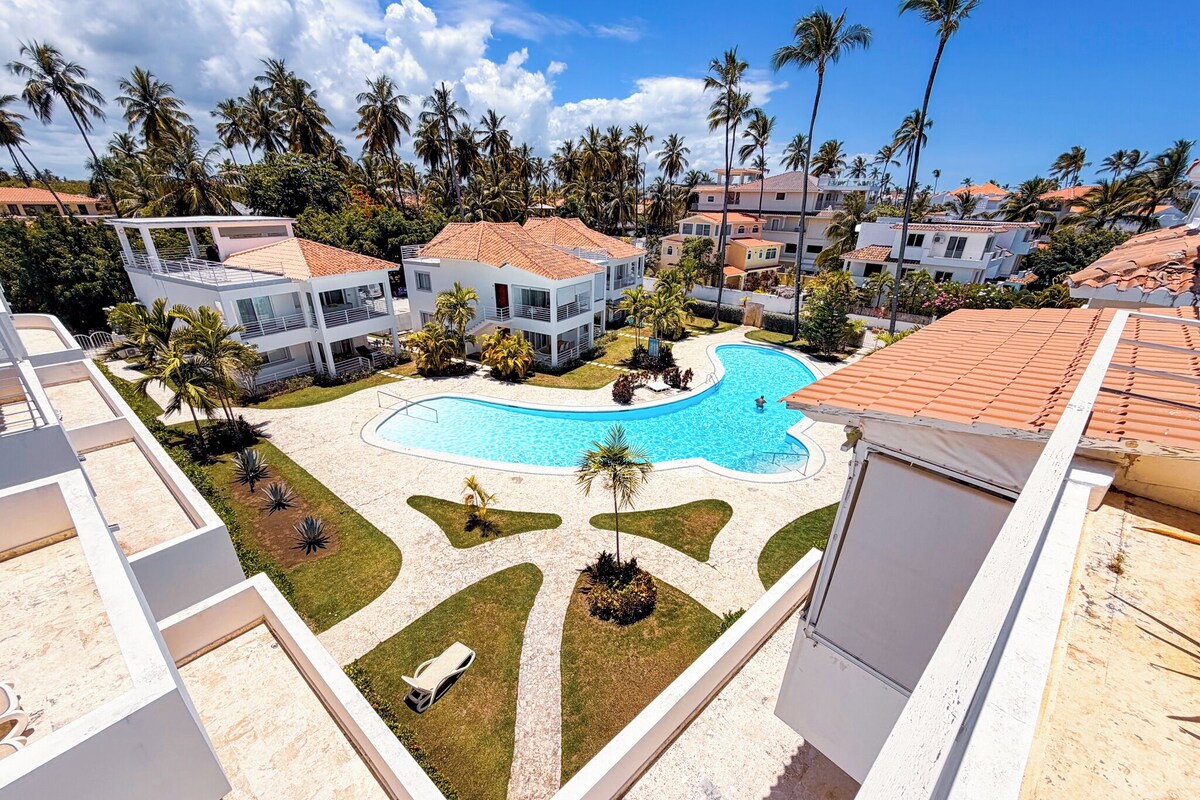 A residential area with a swimming pool surrounded by palm trees and houses with red-tiled roofs.