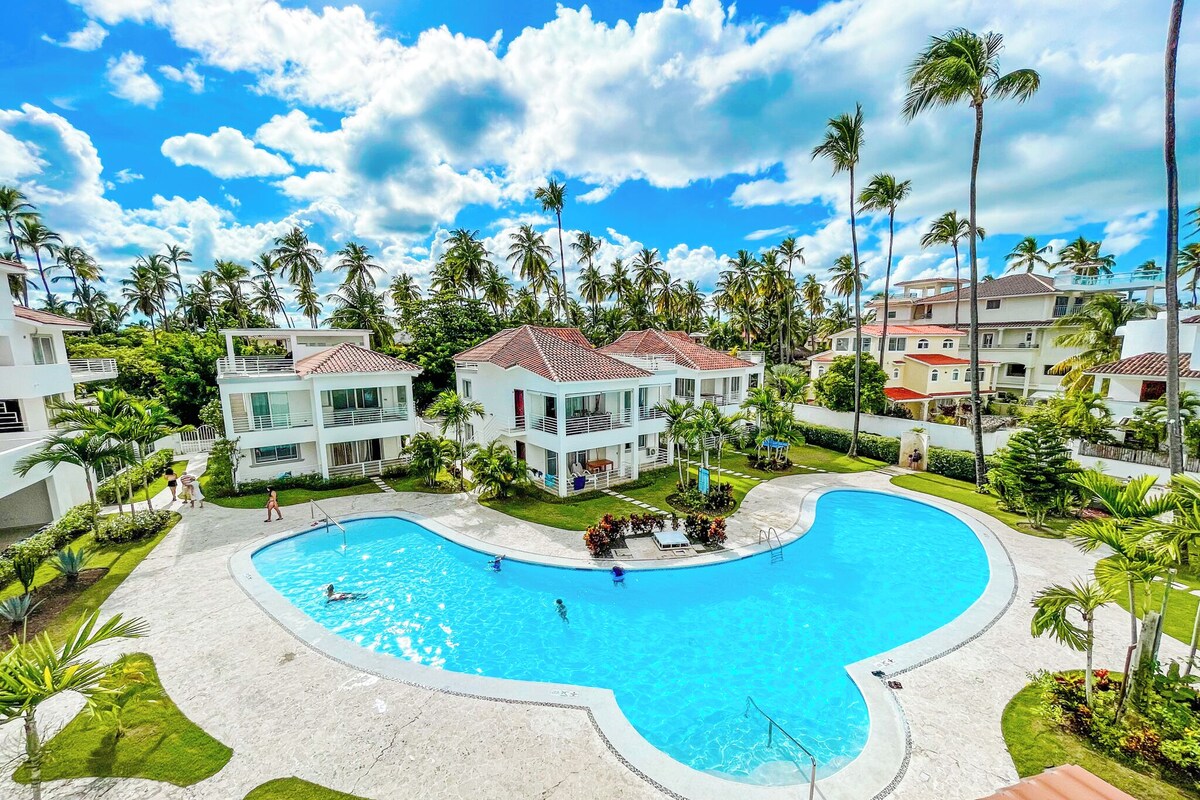 A resort with a large swimming pool, multiple-story buildings, and palm trees.