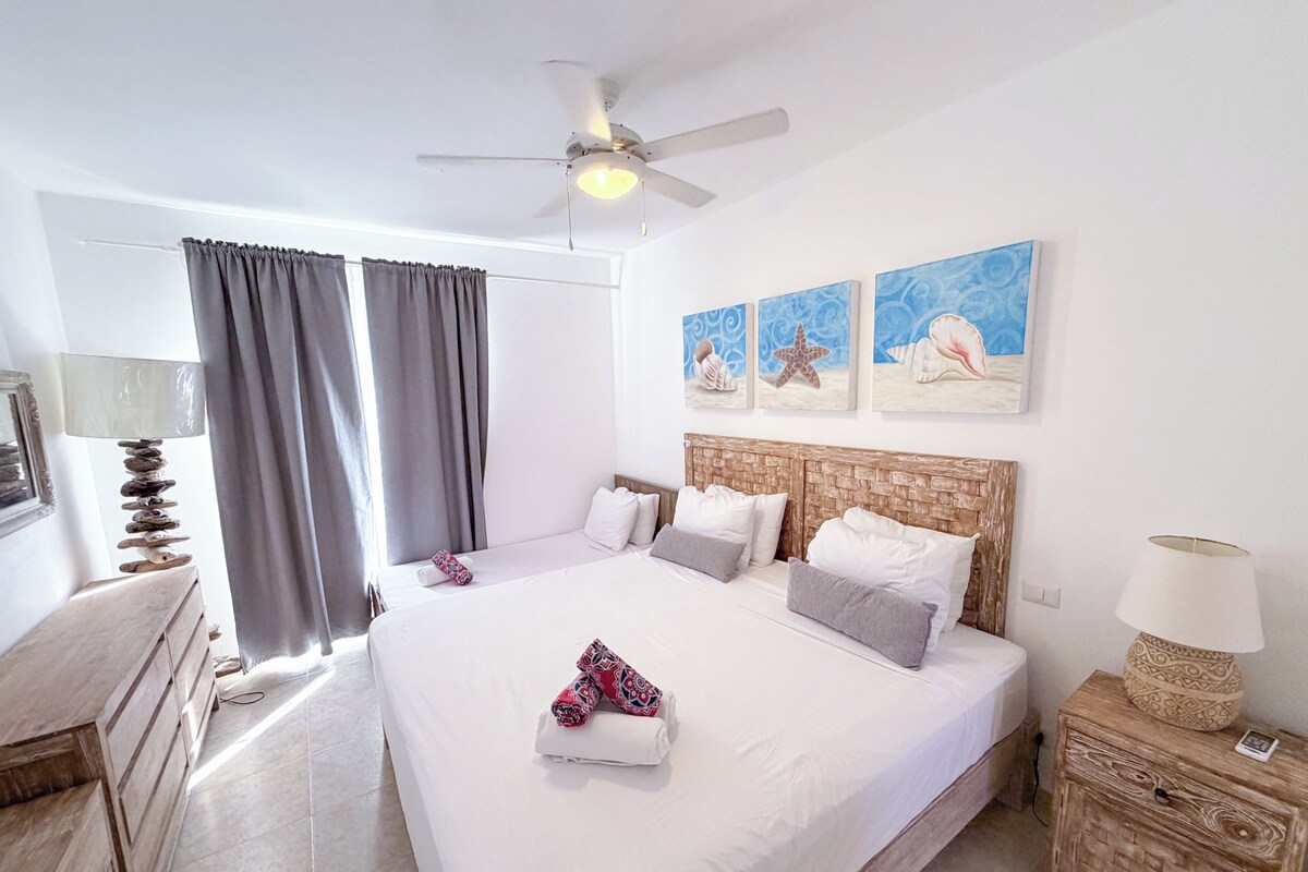 A hotel room with a bed, a ceiling fan, a nightstand, a lamp, and artwork on the wall.