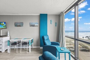 Quarto com uma grande janela com vista para a praia, uma escrivaninha com cadeiras e uma poltrona azul.