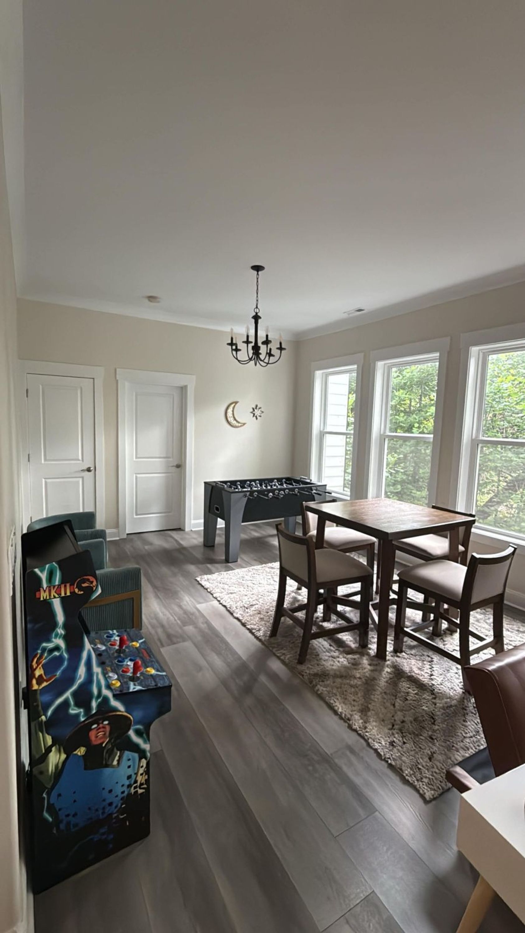 A spacious room with a foosball table, a dining table with chairs, a chandelier, and large windows.