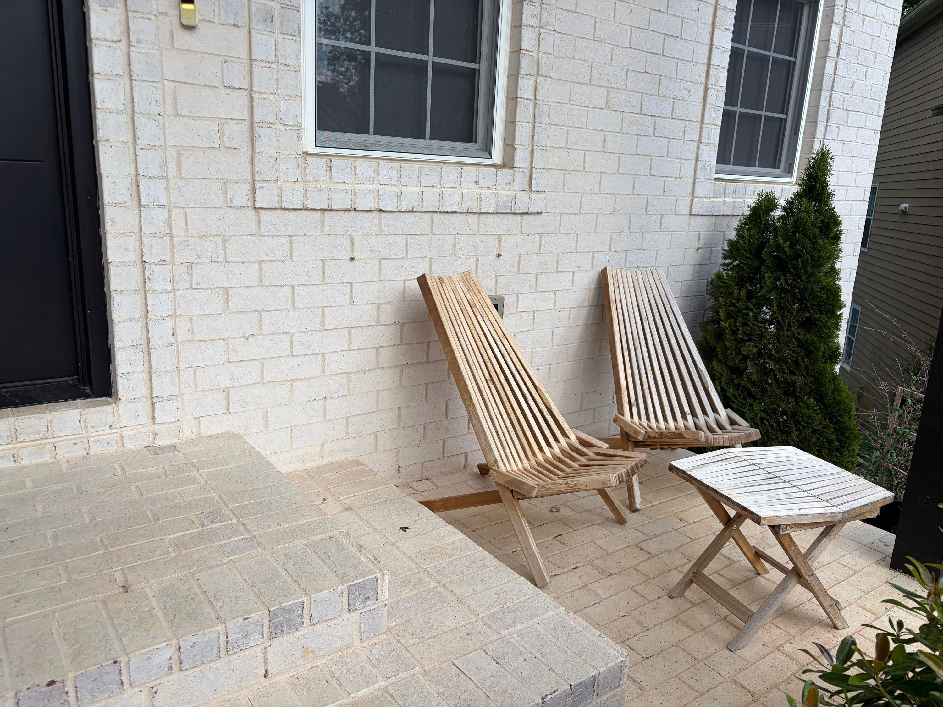 A patio with two wooden lounge chairs and a small table, a white brick wall, and a window.