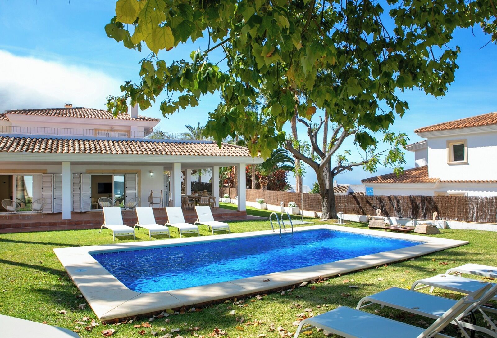 A rectangular swimming pool with a tiled deck, surrounded by white lounge chairs and a covered patio area.