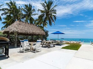 Eine Terrasse am Strand mit einer Struktur unter einem Reetdach, weißen Liegestühlen, einem blauen Sonnenschirm und Blick aufs Meer.