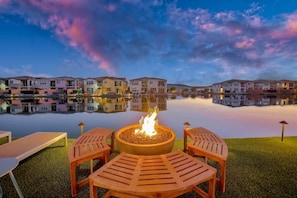 A lakeside outdoor seating area with a fire pit, lounge chairs, and a row of buildings in the background. - Sunset by the Lake | Heated Pool, Spa & Arcade (Indio)
