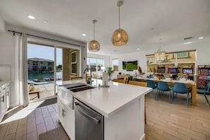 A modern kitchen with a large island, dining area with blue chairs, and a view of a pool. - Sunset by the Lake | Heated Pool, Spa & Arcade (Indio)
