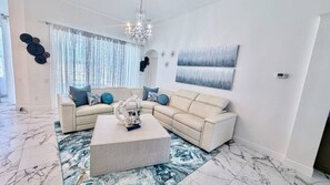 A living room with a sectional sofa, a coffee table, a chandelier, and a large window with curtains.