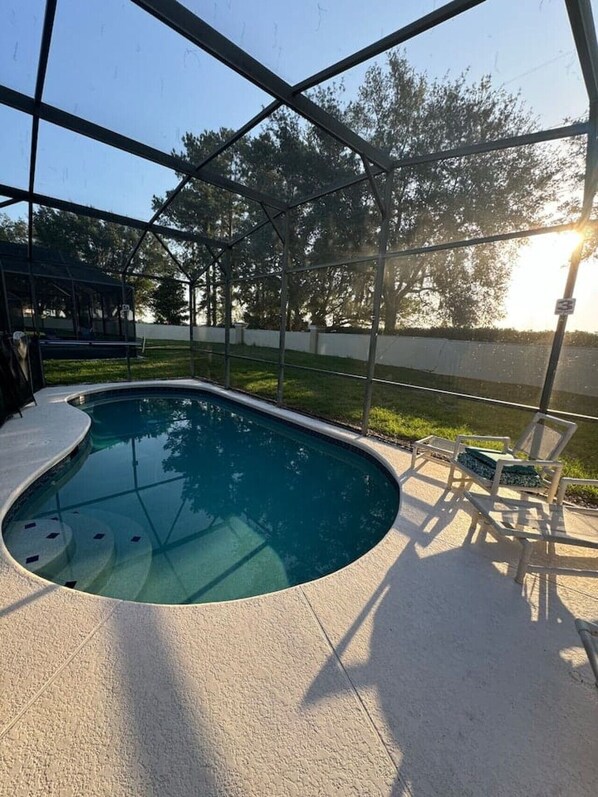 A swimming pool with a curved edge, surrounded by a covered area with chairs and a view of trees.