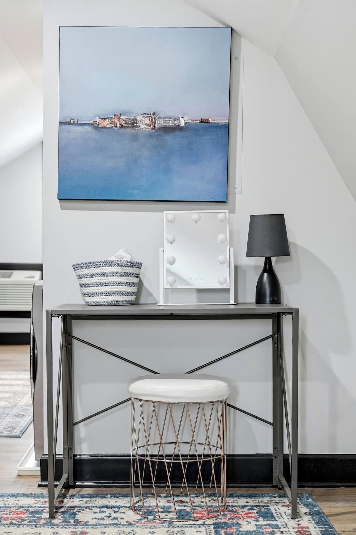 Ett rum med ett vitt skrivbord, en vit stol, en svart lampa, en vit sminkbord med belysning och en målning som föreställer ett kustlandskap.