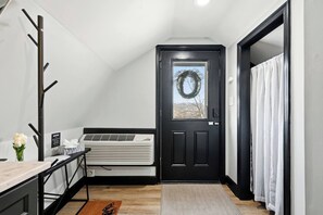 Une pièce avec une porte noire, un climatiseur blanc, une petite table sur laquelle est posé un vase de fleurs et un porte-manteau.