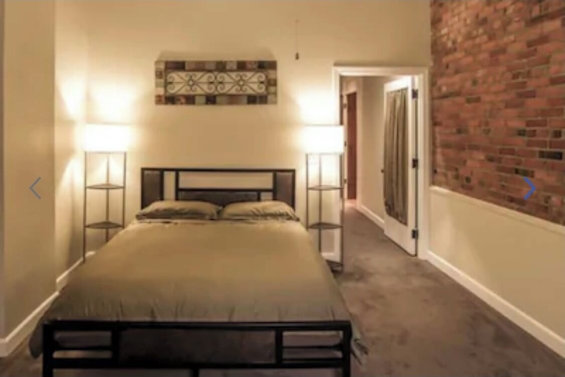 A narrow hallway with a bed at the end, a brick wall on the right, and a door leading to another room.